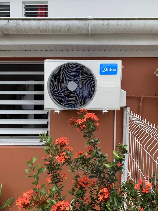Façade extérieure d'habitation avec climatiseur Midea installé sur mur, plantes ornementales rouges au premier plan