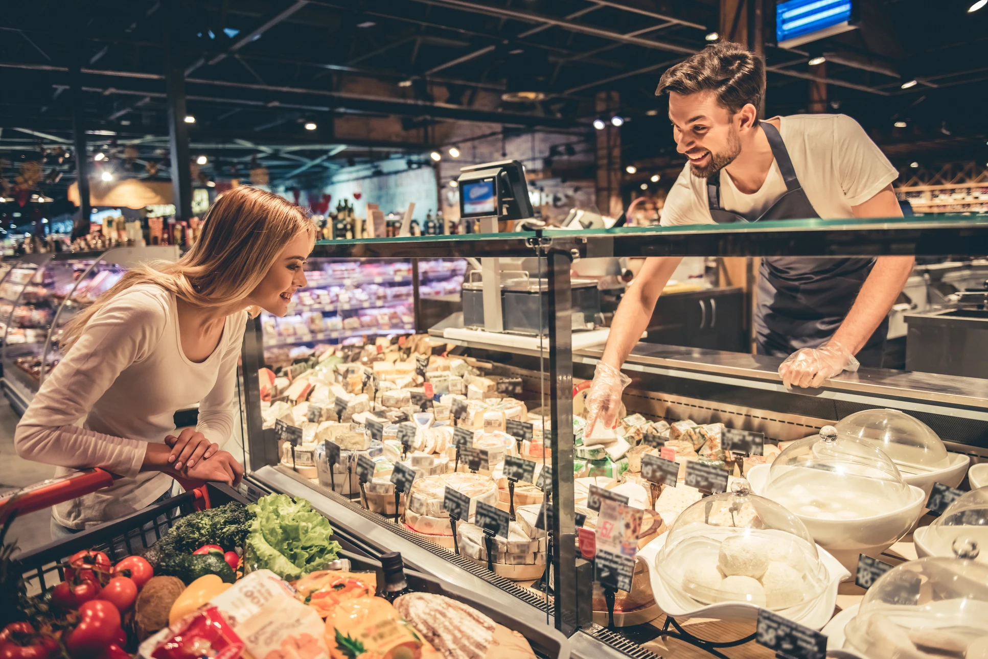 Une femme cliente examine les produits d'un comptoir réfrigéré de fromages dans un supermarché moderne, tandis qu'un employé en tablier noir lui présente une sélection